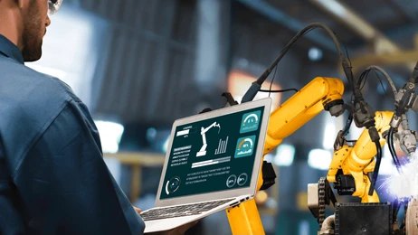 A technician using a laptop to monitor and control a yellow robotic arm in an industrial automation setting.