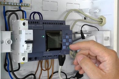 A finger pressing a button on a PLC module mounted in a control panel.