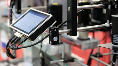 Industrial touchscreen control panel mounted on machinery, displaying operational data with connected cables.